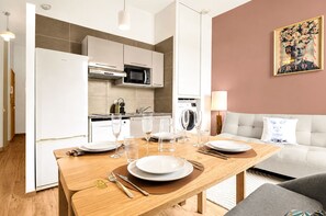 Eine moderne Küche mit weißen Geräten, ein Essbereich mit einem Holztisch für vier Personen und ein Wohnzimmer mit einer Couch und einem gerahmten Bild an der Wand.