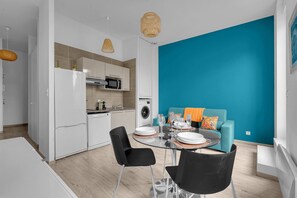 A modern kitchen with white appliances, a dining area with a round table and chairs, and a teal-colored sofa with colorful cushions. - Terrasse Riquet - en plein coeur de Toulouse (Toulouse)