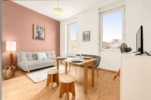 A modern living room with a sofa, dining table, and chairs, a large window with a city view, and a flat-screen TV on a cabinet. - Terrasse Riquet - en plein coeur de Toulouse (Toulouse)