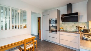 A modern kitchen with white cabinets, a built-in oven, and a microwave. There is a dining table with chairs and a window with blinds. - Ty Bigorn'oh - Three Bedroom House, Sleeps 7 (Benodet)