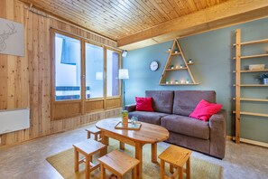 Una habitación acogedora con paredes y techo de madera, un sofá marrón con cojines rojos, una mesa de madera con bancos y una ventana con vistas.