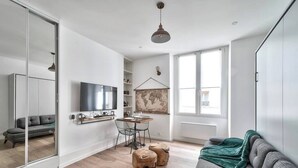 A modern living room with a grey sofa, wooden stools, a dining table, and a TV. - Magnifique studio - Le Marais (Paris)