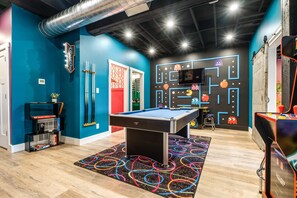 A pool table in a room with blue walls, a neon sign, and a wall with arcade game graphics.
