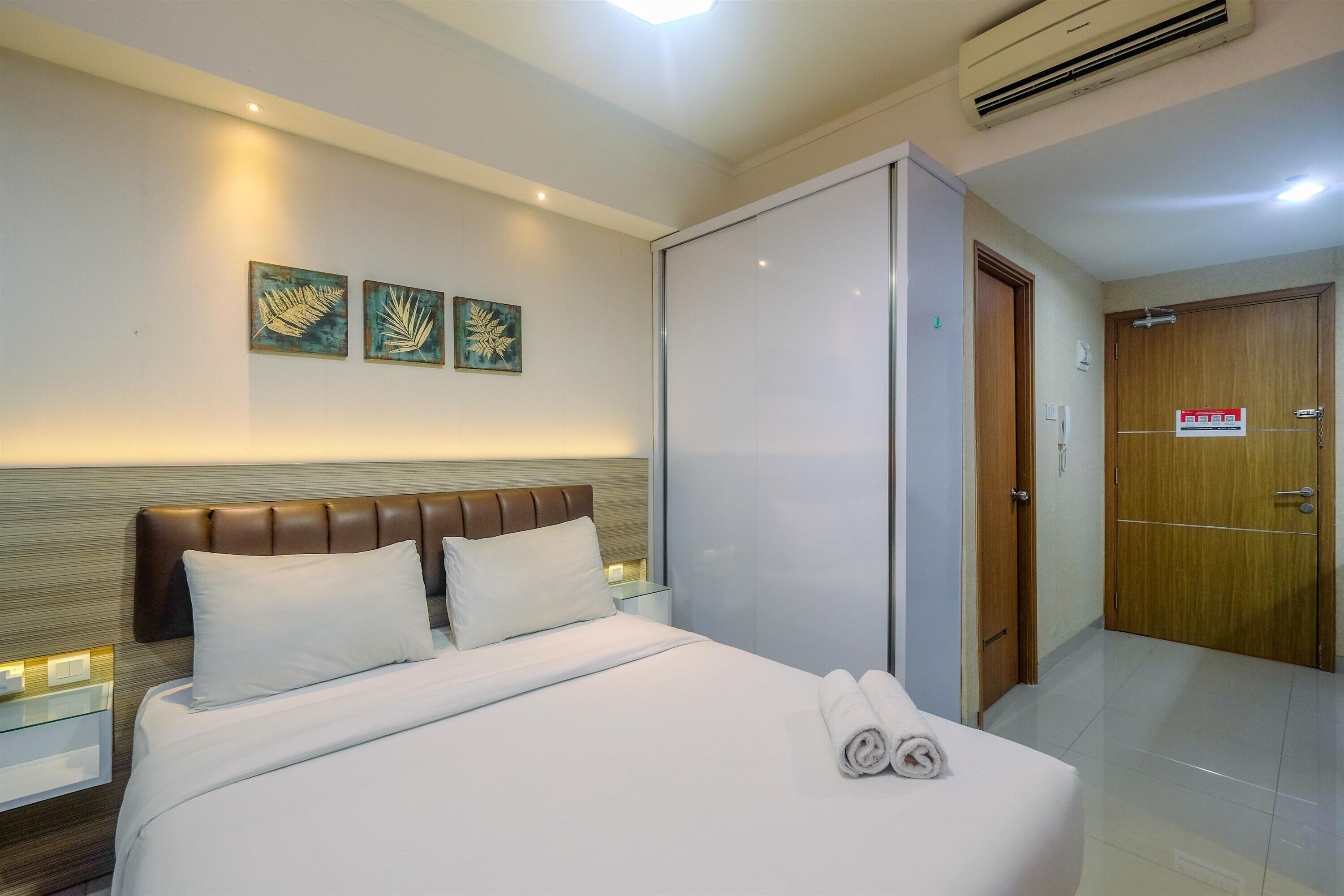 A hotel room with a bed, two pillows, a nightstand, a wall-mounted artwork, an air conditioning unit, and a wardrobe.