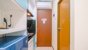 A narrow hallway with a kitchen area, a wooden door, and a blue trash bin. - Homey And Cozy Living Studio Room At Tifolia Apartment (Jakarta)