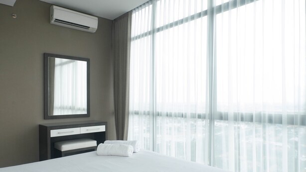 Une chambre d’hôtel avec un grand lit, une table de chevet avec un miroir, un banc et une vue sur le paysage urbain à travers des rideaux transparents.