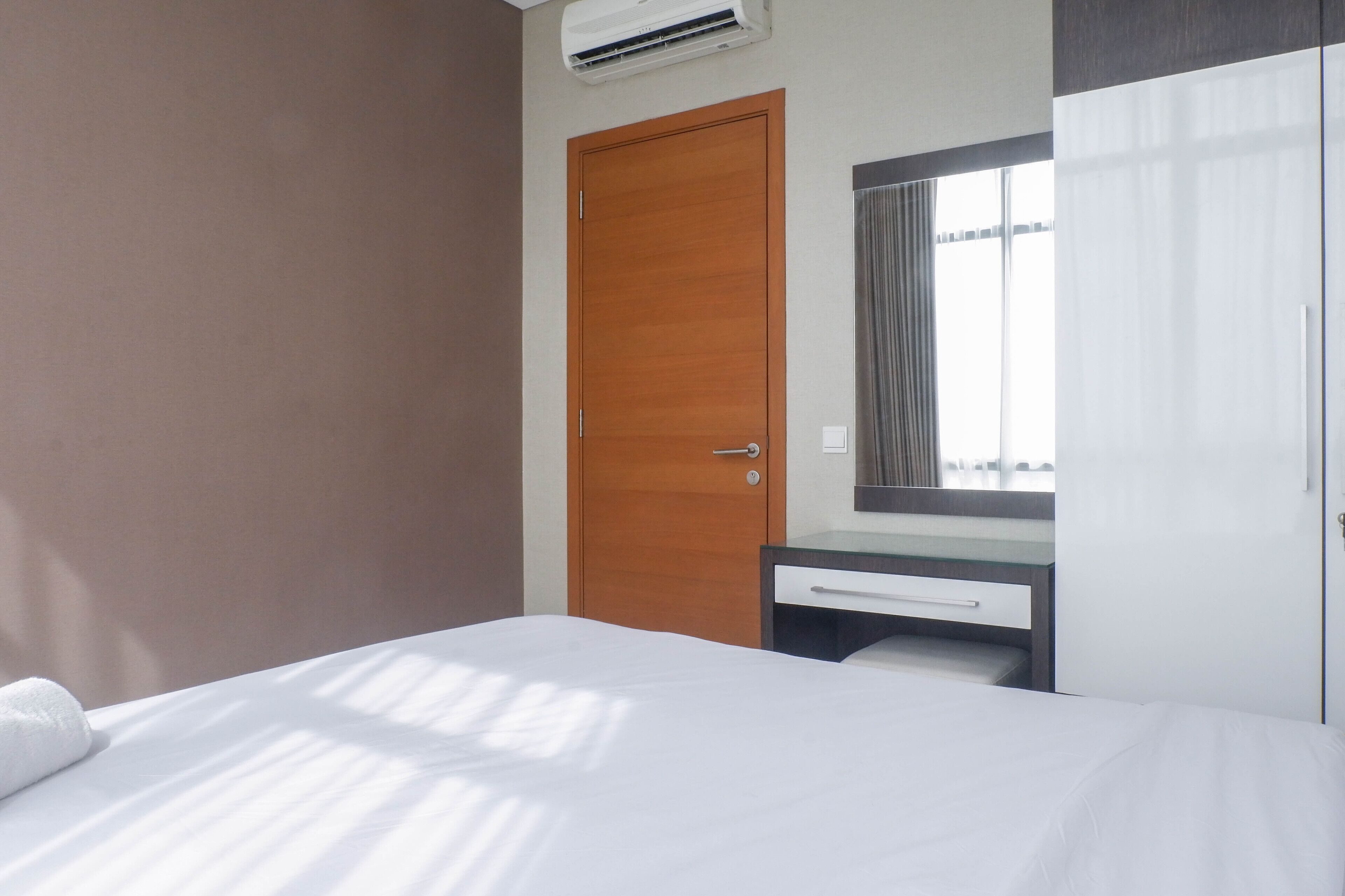A hotel room with a bed, a wooden door, a small desk with a mirror, and a window with curtains.