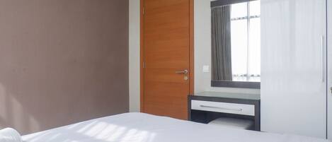 A hotel room with a bed, a wooden door, a small desk with a mirror, and a window with curtains.