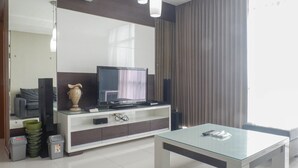 Un salon moderne avec une table basse en verre, un téléviseur à écran plat posé sur un meuble de télévision blanc, un vase et une grande fenêtre avec des rideaux.