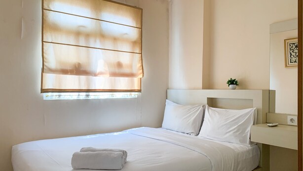 A neatly made bed with white linens and a rolled towel, a bedside table with a small plant, and a window with sheer curtains. - Modern Look And Comfortable 2Br Green Pramuka City Apartment (Jakarta)