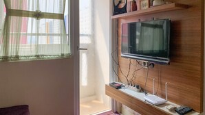 A living room with a TV mounted on a wooden wall, a window with sheer curtains, and a small shelf with decorative items. - Modern Look And Comfortable 2Br Green Pramuka City Apartment (Jakarta)