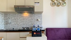 A compact kitchenette with a gas stove, a microwave, and a tiled backsplash. There are two bottles of water on a box labeled 'Travelio Property Management'. A purple couch is visible to the right. - Modern Look And Comfortable 2Br Green Pramuka City Apartment (Jakarta)
