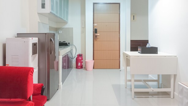 Cozinha compacta com geladeira, fogão e pia. Uma porta de madeira dá acesso a outro cômodo. Uma mesa branca com um objeto preto é visível.
