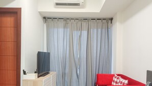 Um espaço de estar compacto com um sofá vermelho, um pequeno armário de madeira, uma televisão e uma janela com cortinas azul-claras.