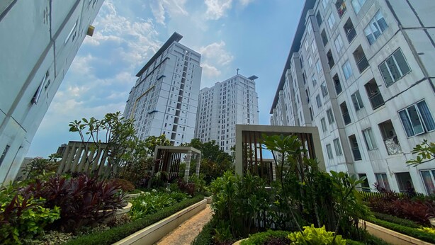 A modern residential building complex with multiple high-rise structures, a well-maintained garden area with various plants, and a clear sky. - Modern Design And Cozy Studio At Bassura City Apartment (Jakarta)