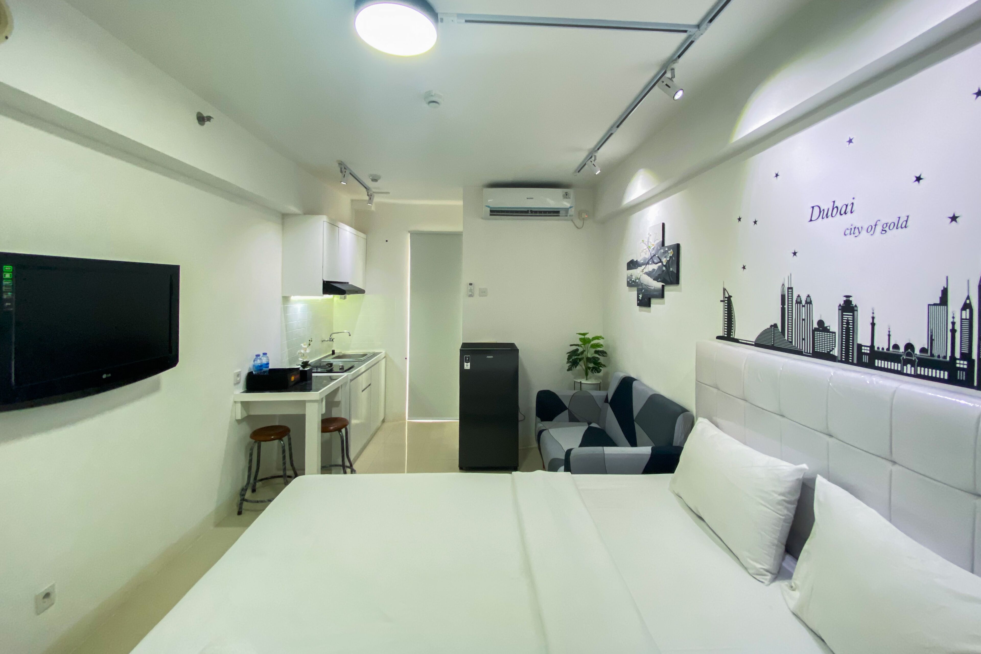 Une chambre d’hôtel compacte comprenant un lit, une petite kitchenette, un téléviseur et une fresque murale représentant le paysage urbain de Dubaï.