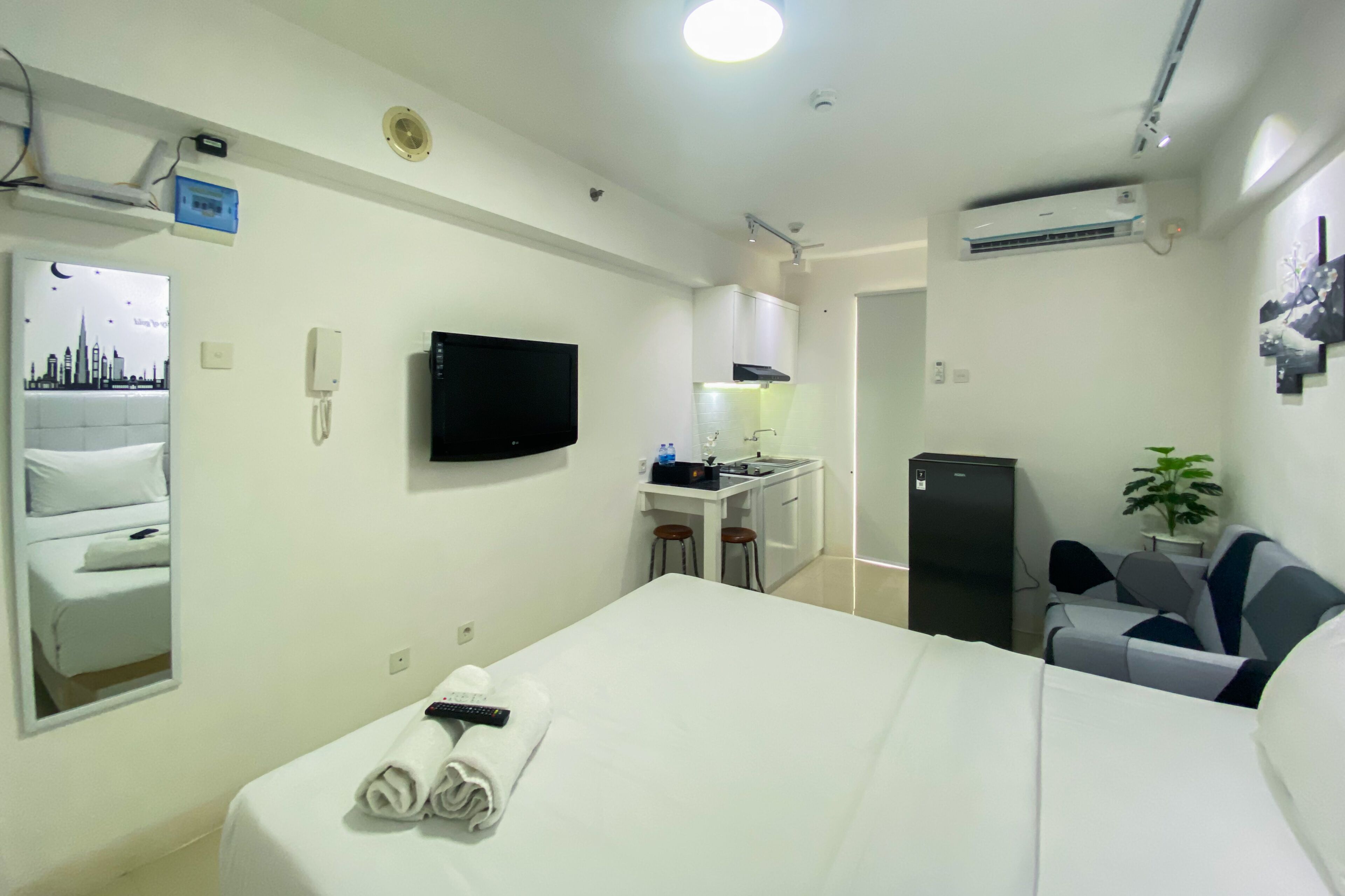 A hotel room with a bed, a TV, a desk with a chair, a small kitchenette, a refrigerator, and a plant.