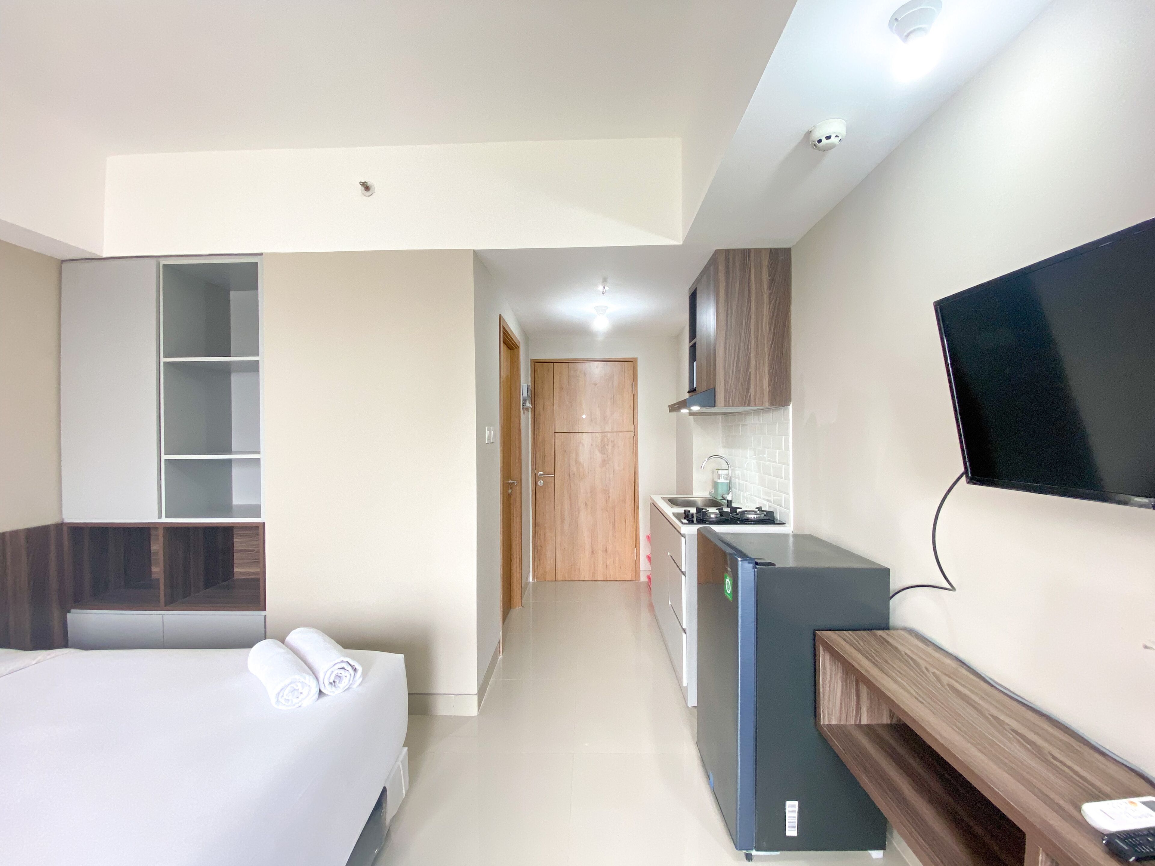 A compact hotel room with a bed, a small kitchenette, a TV, and a wooden bench.