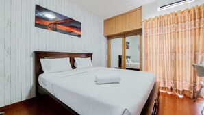 A hotel room with a bed, wooden headboard, white walls, a painting, a window with curtains, and an air conditioning unit. - Good And Clean Studio Room Serpong Greenview Apartment (South Tangerang)