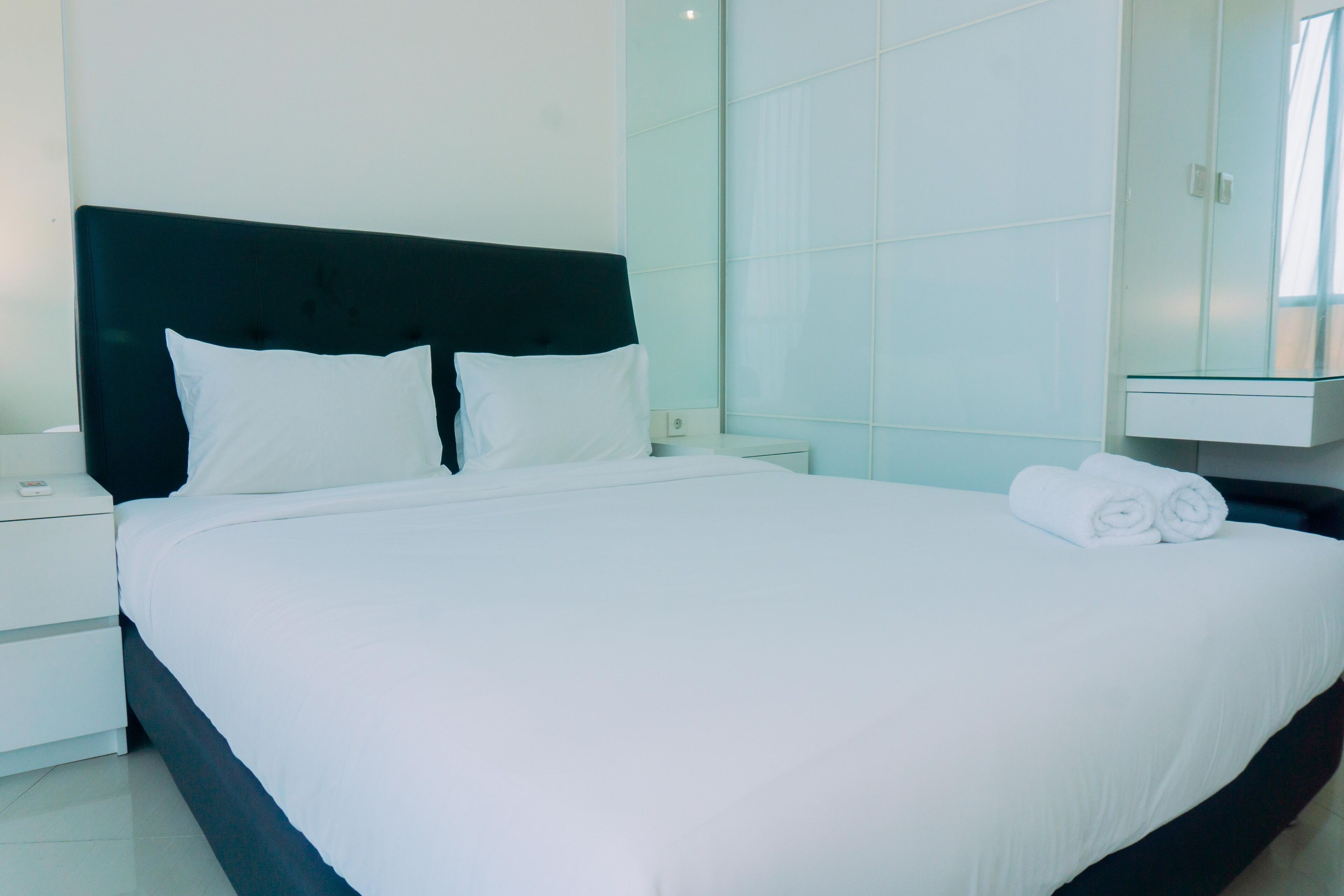 A neatly made bed with white linens and pillows, a bedside table with a lamp, and a glass cabinet with folded towels.