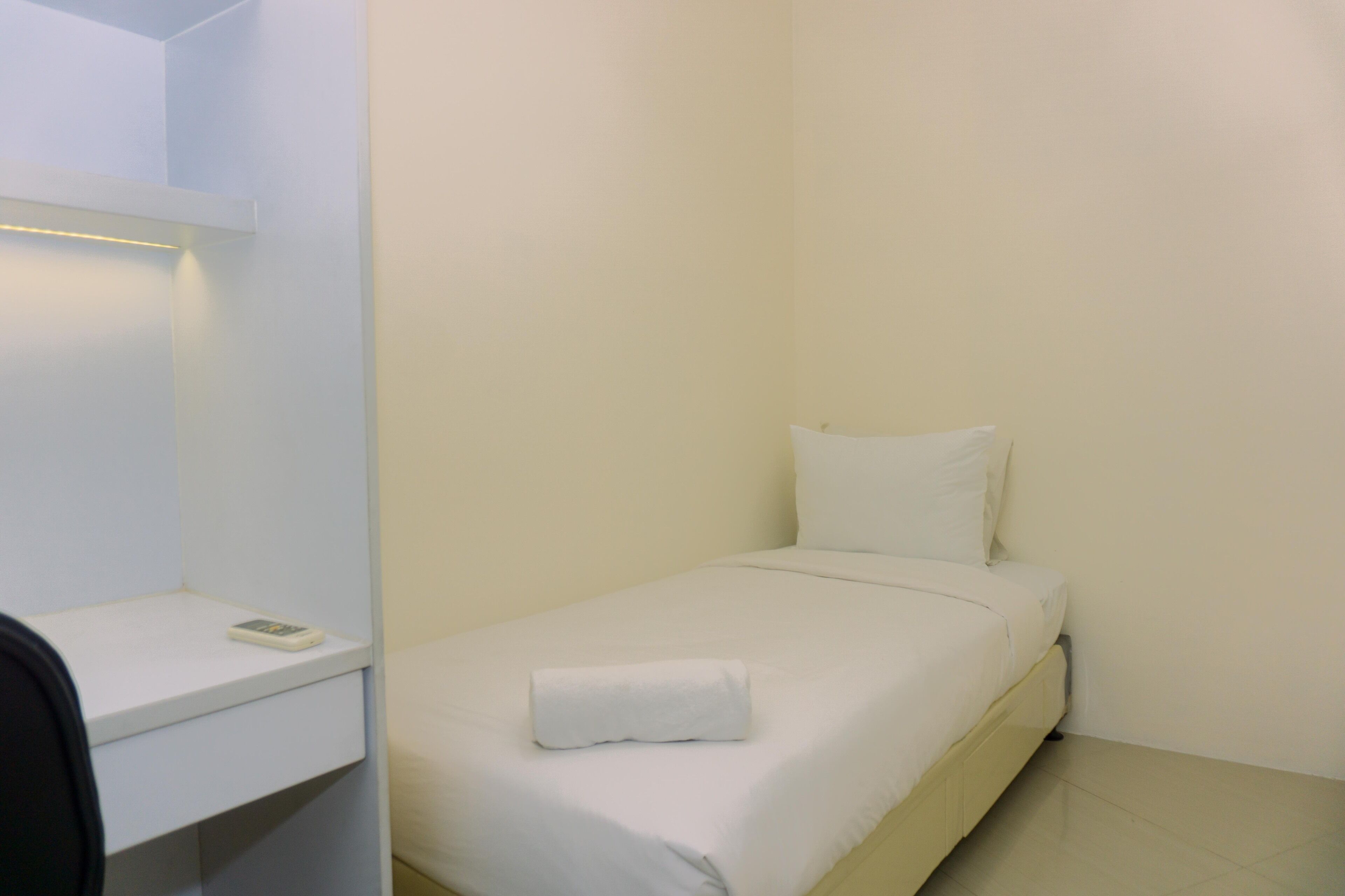 A small, single-bed room with a white bed, a white pillow, and a white blanket. There is a small shelf with a phone on it.