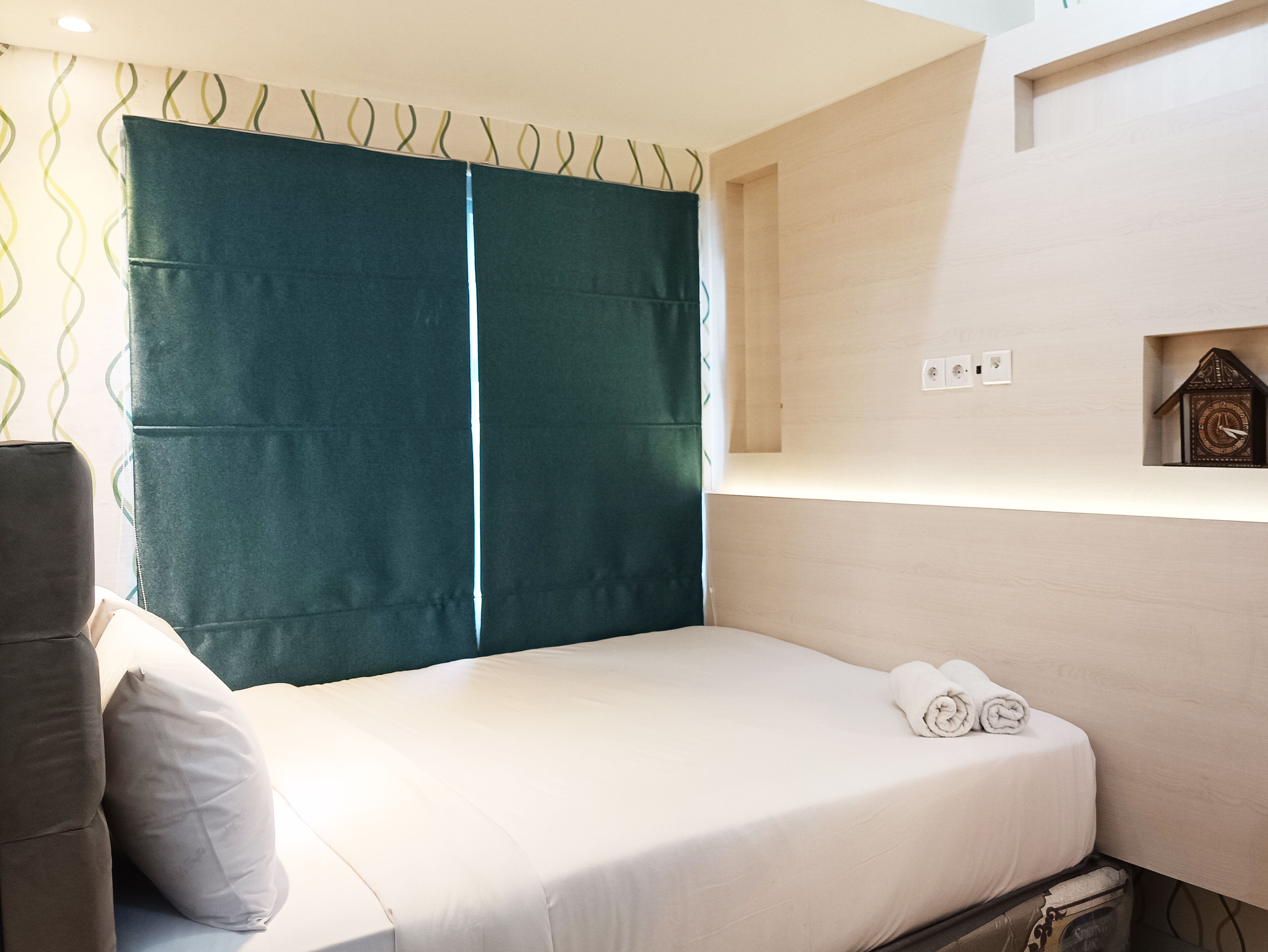 Een hotelkamer met een bed, een groen gordijn, een klein horloge aan de muur en een handdoek op het bed.