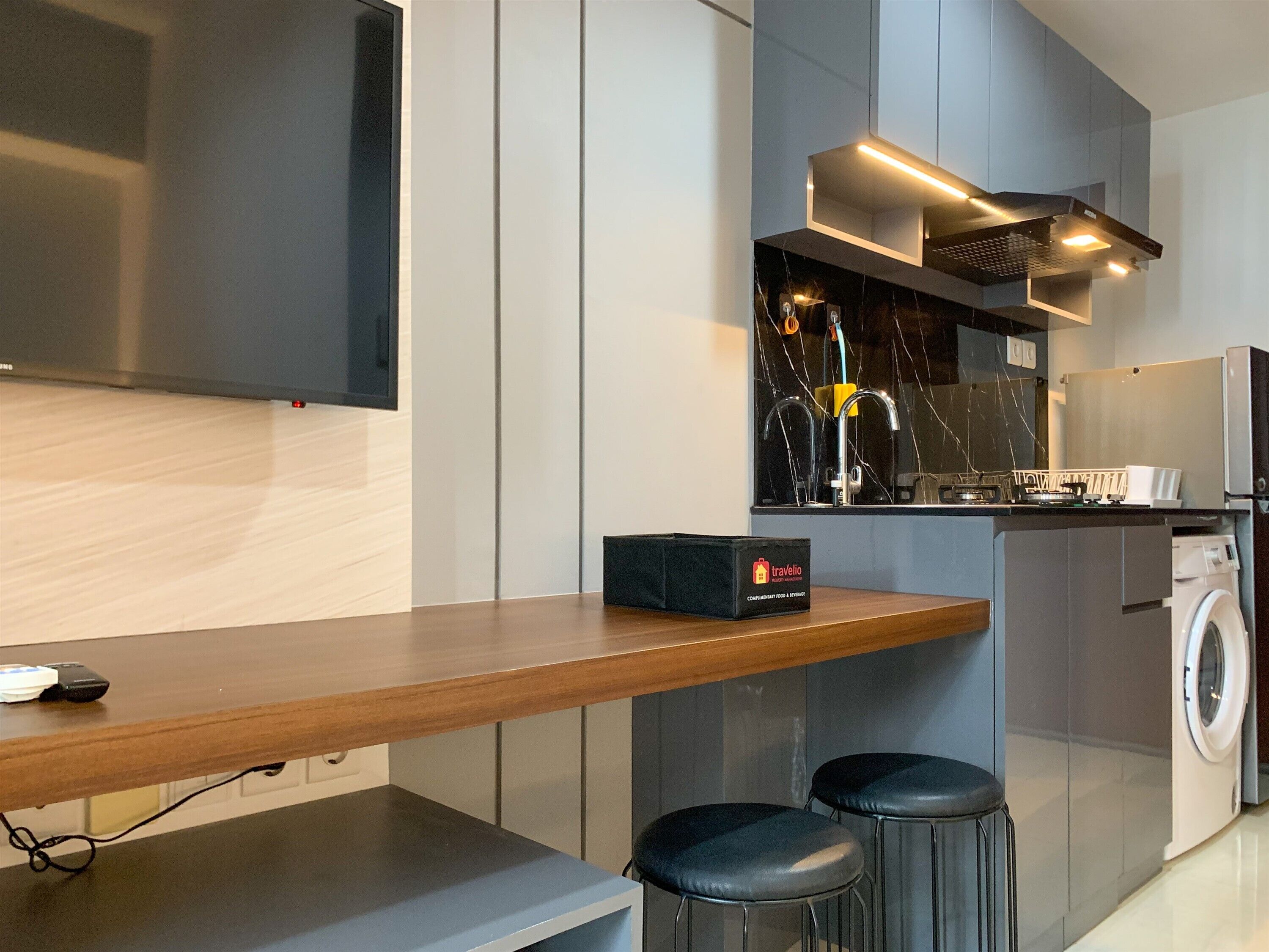 A modern kitchen with a wooden countertop, two bar stools, a washing machine, and a travel bag on the counter.
