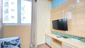 A hotel room with a bed, a TV mounted on the wall, a window with a curtain, and a view of buildings outside. - Modest 2Br Apartment At Suites @Metro (Bandung)