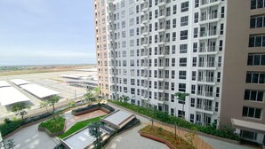 A high-rise residential building with multiple balconies, a landscaped courtyard, and a view of a distant airport. - Simply And Cozy Living Studio At Tokyo Riverside Pik 2 Apartment (Teluknaga)