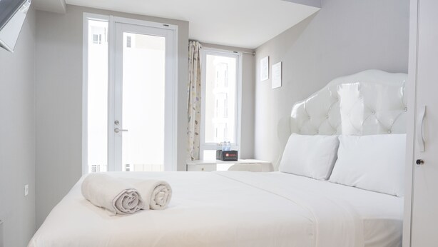 A hotel room with a large bed, a bedside table, a window with curtains, and a door. - Simply And Cozy Living Studio At Tokyo Riverside Pik 2 Apartment (Teluknaga)
