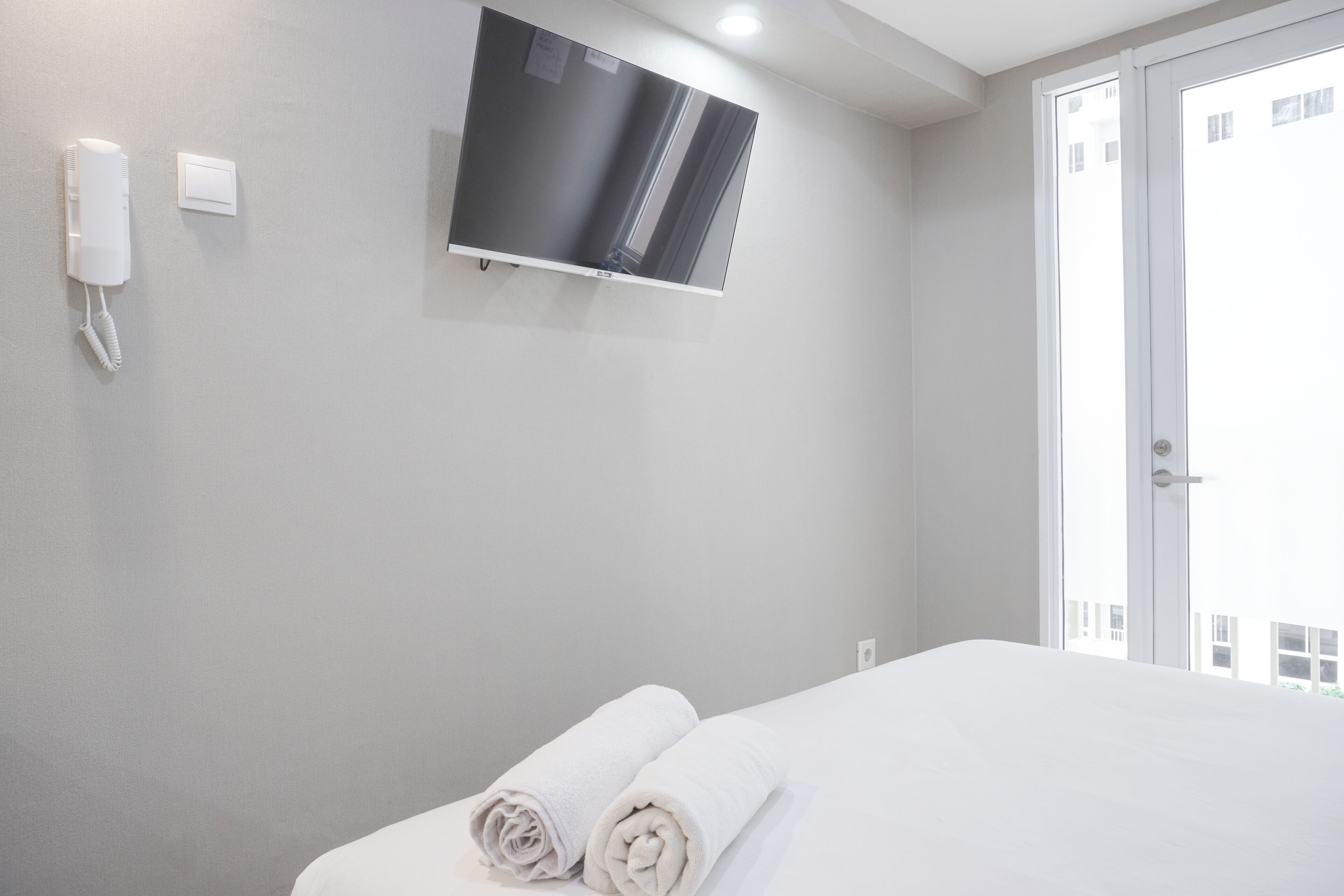 A hotel room with a bed, a TV mounted on the wall, a phone, and a door with a view of buildings outside.
