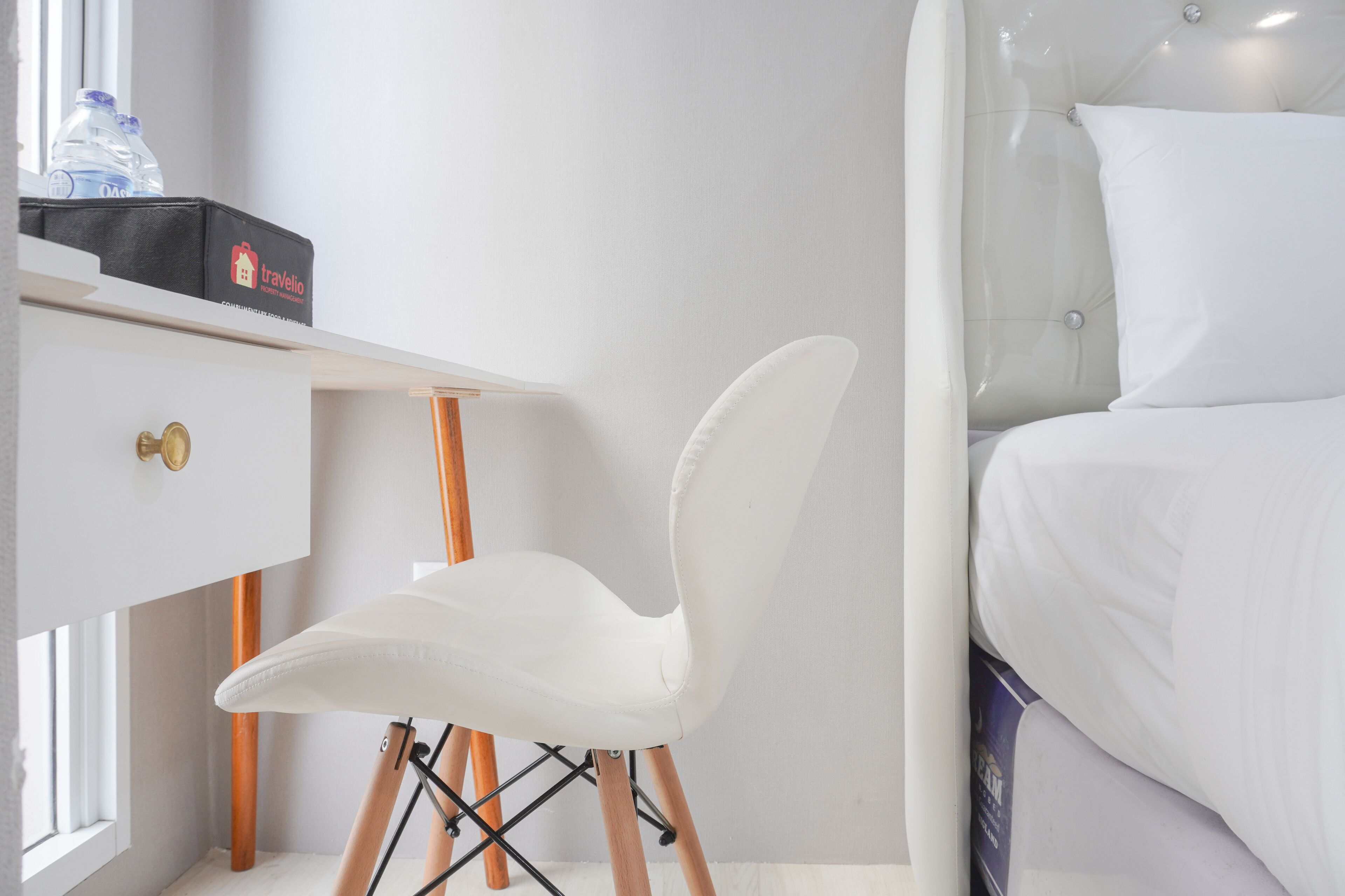 A modern hotel room with a white desk, a white chair, a bed with white bedding, and a window with natural light.