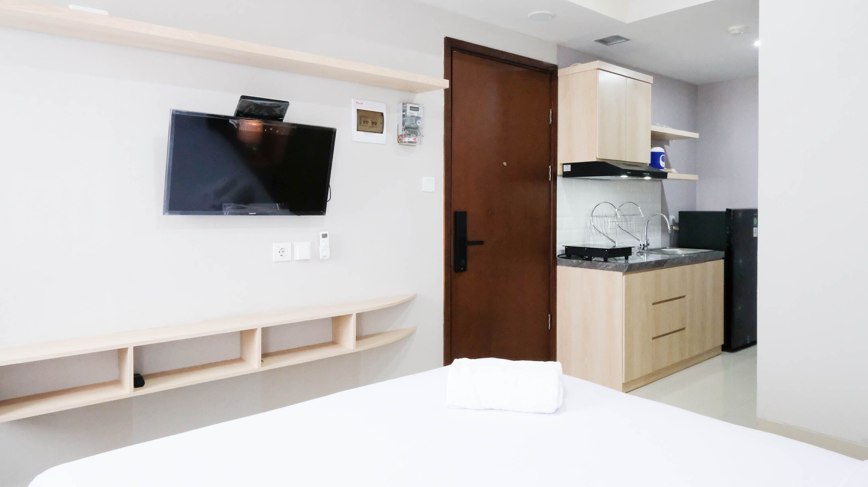 A modern hotel room with a flat-screen TV mounted on the wall, a wooden door, a kitchen area with a sink and cabinets, and a white bed with a towel.