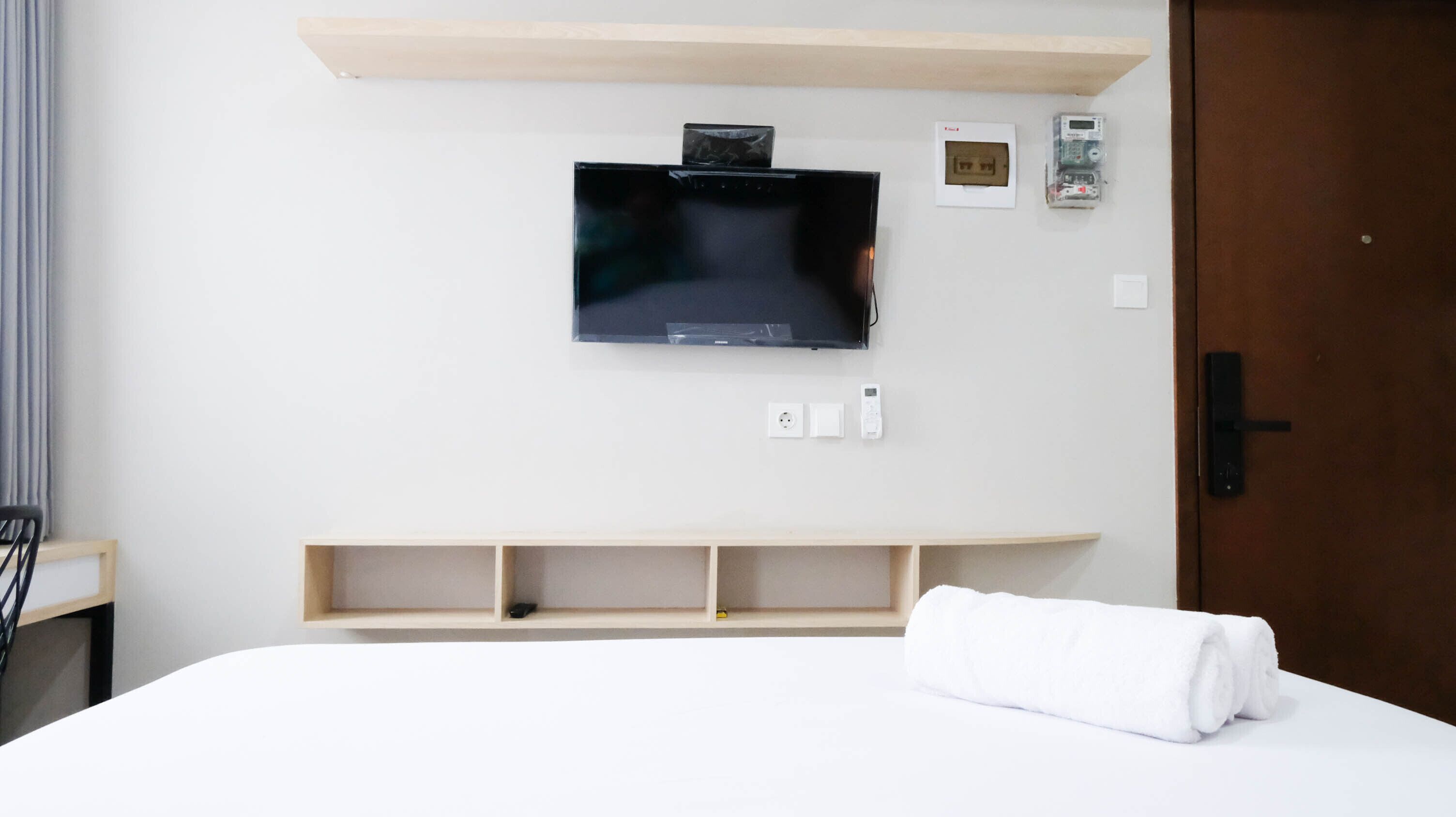 Ett hotellrum med en säng, en väggmonterad TV, en hylla i trä och en dörr.