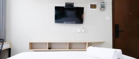 A hotel room with a bed, a mounted TV, a wooden shelf, and a door.