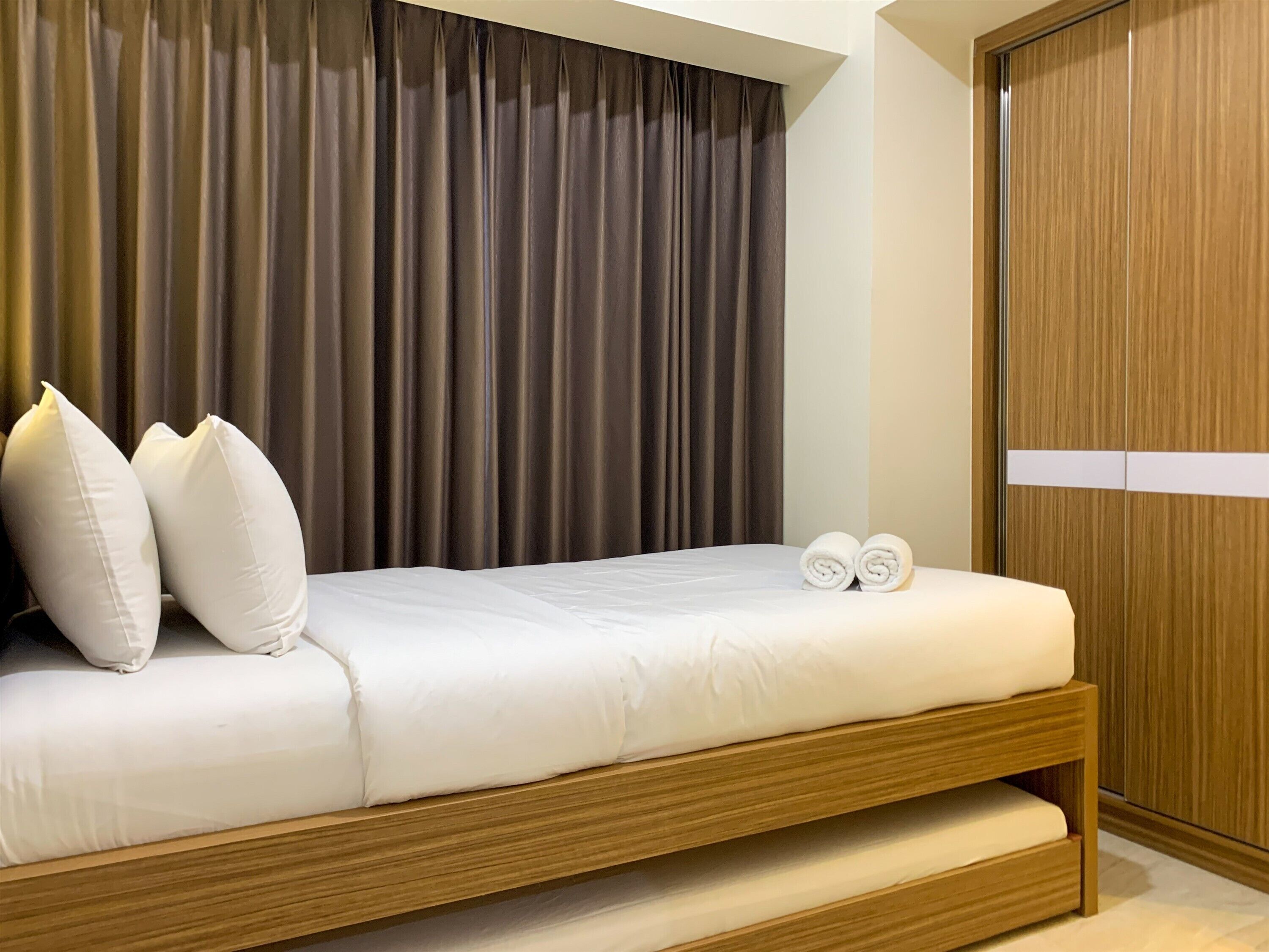A neatly made bed with white linens and pillows, a wooden headboard, and a pair of rolled towels on the side.