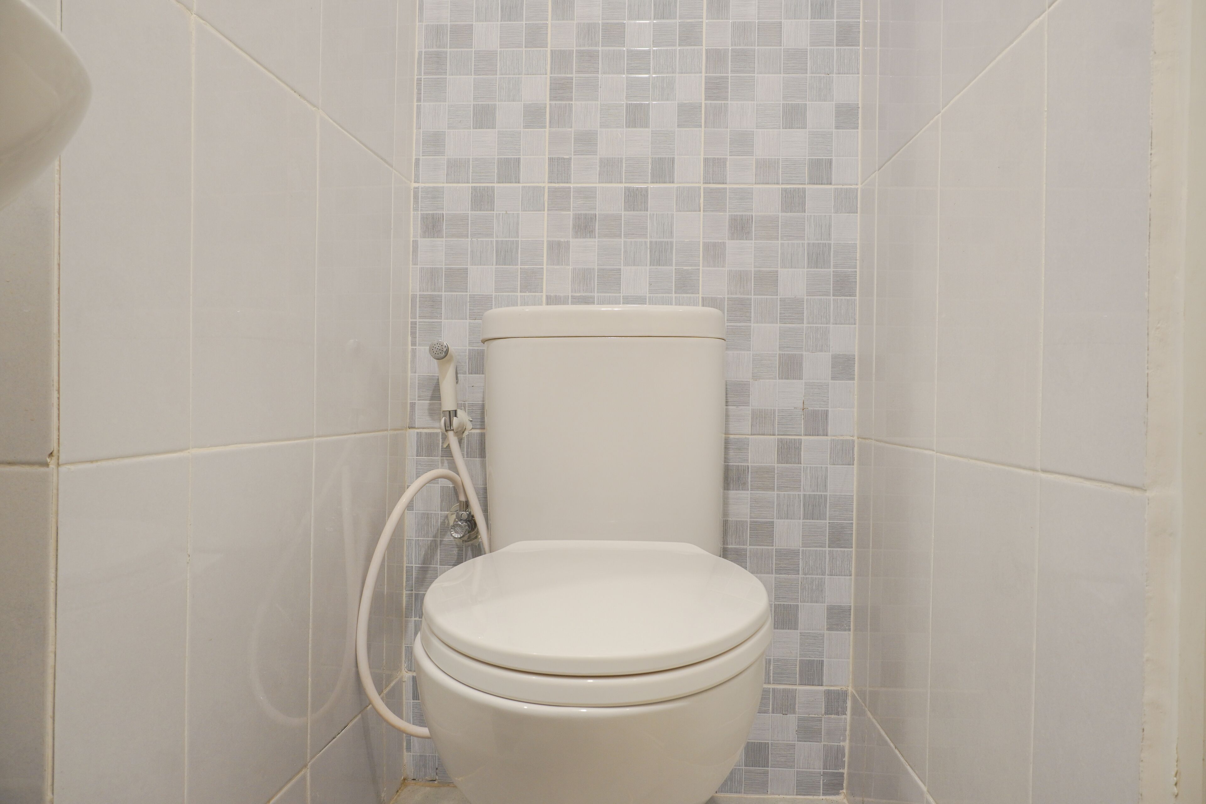 A toilet with a white seat and lid, set against a wall with small, light-colored tiles.