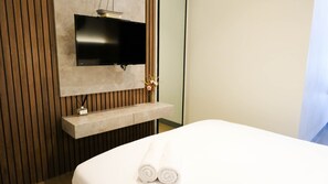 A hotel room with a bed, a flat-screen TV mounted on a wooden panel wall, a small shelf with a bottle, and a vase with dried flowers. - Aesthetic 2Br Connected To Mall At Supermall Mansion Apartment (Surabaya)