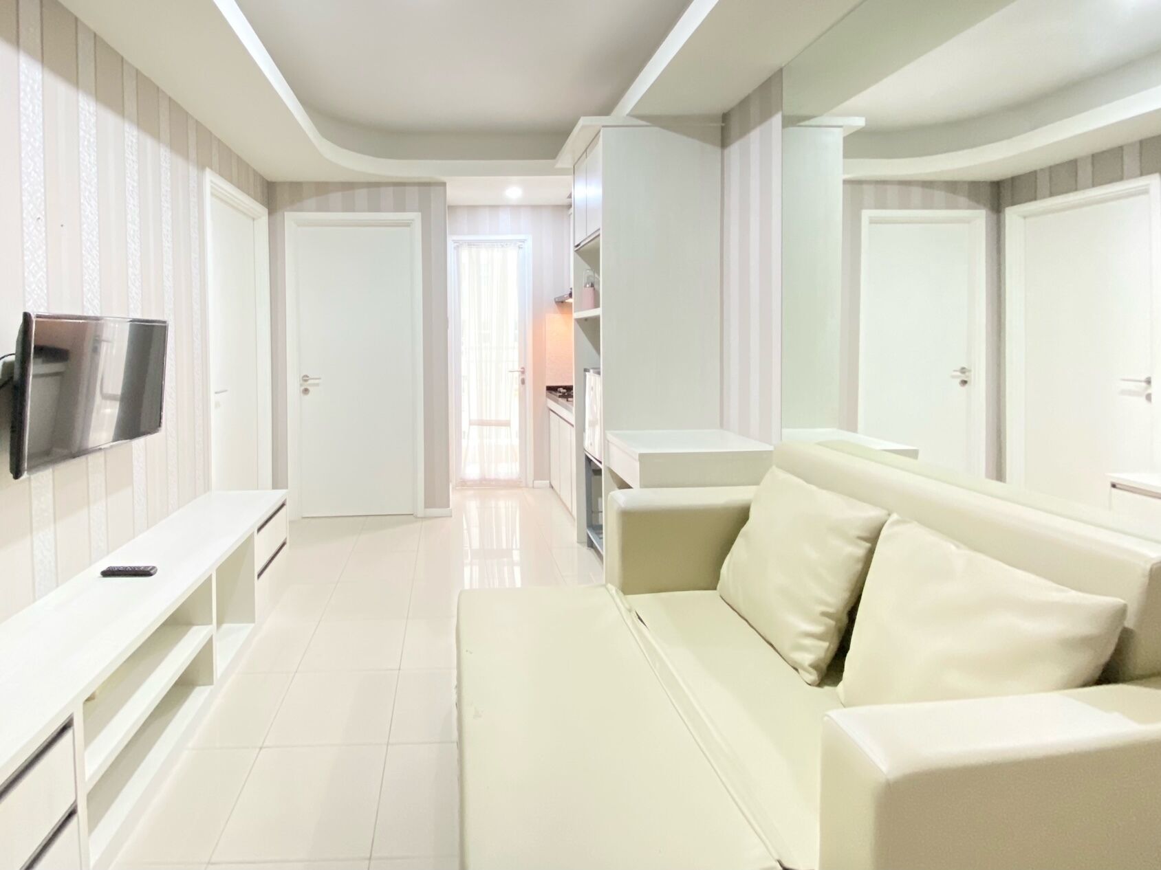A modern, well-lit living room with a flat-screen TV mounted on the wall, a beige sofa with cushions, and a white entertainment unit.