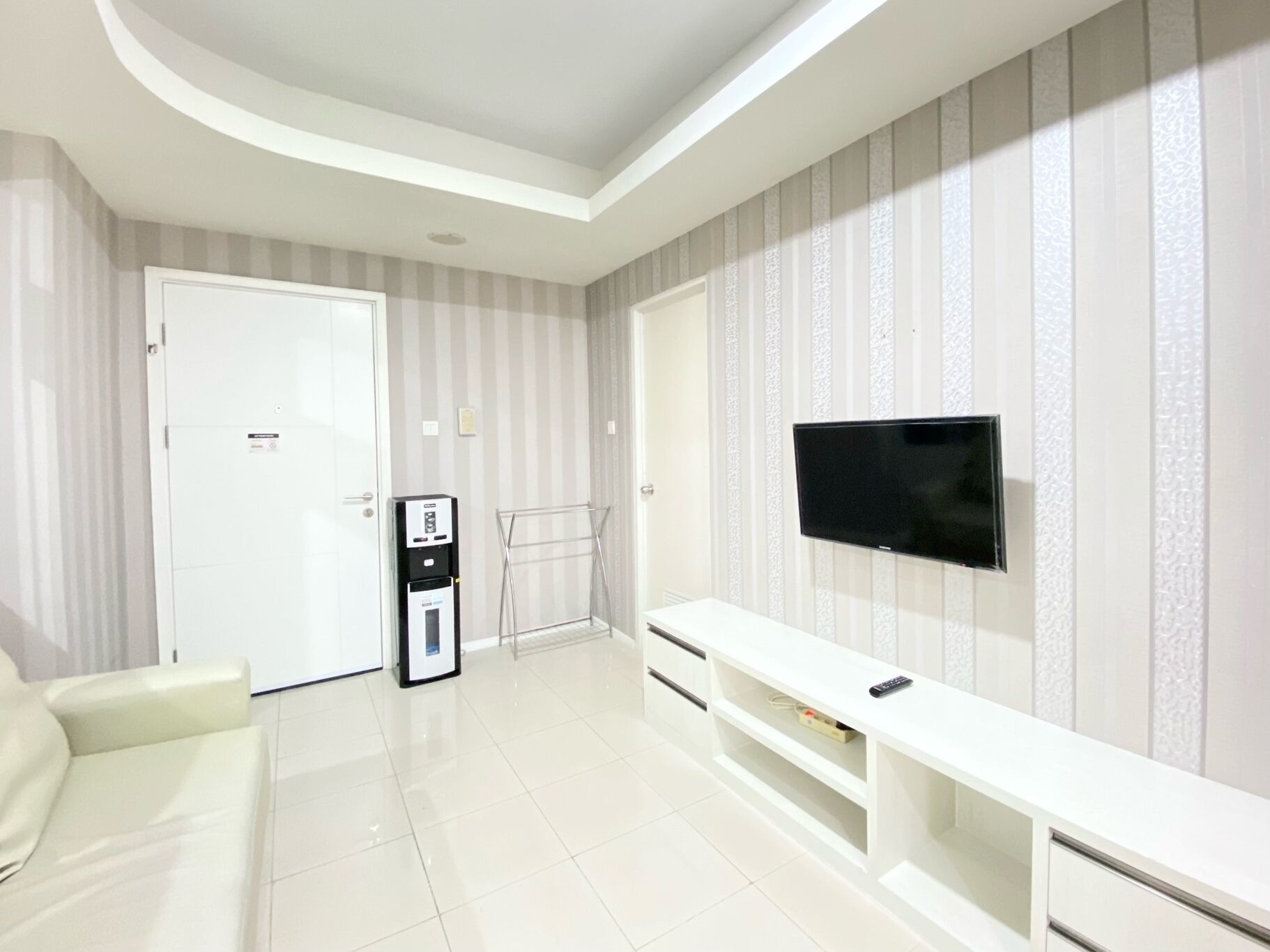 Un salon moderne avec un canapé blanc, un téléviseur à écran plat fixé au mur, un meuble de rangement blanc et un distributeur d’eau.