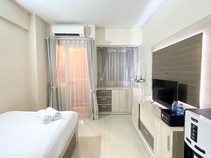 A hotel room with a bed, a desk with a flat-screen TV, an air conditioning unit, and a minibar. - Comfortable Studio Apartment For 1 Pax Grand Sentraland Karawang (Karawang)