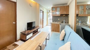A modern living room with a sofa, TV, and wooden coffee table. A kitchen with wooden cabinets and a tiled backsplash is visible in the background. - Nice 2Br Apartment At Gateway Ahmad Yani Cicadas (Bandung)