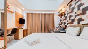 A hotel room with a bed, a desk with a chair, a television, and a wall with a geometric pattern. - Warm And Homey Studio Room Casa De Parco Apartment (South Tangerang)