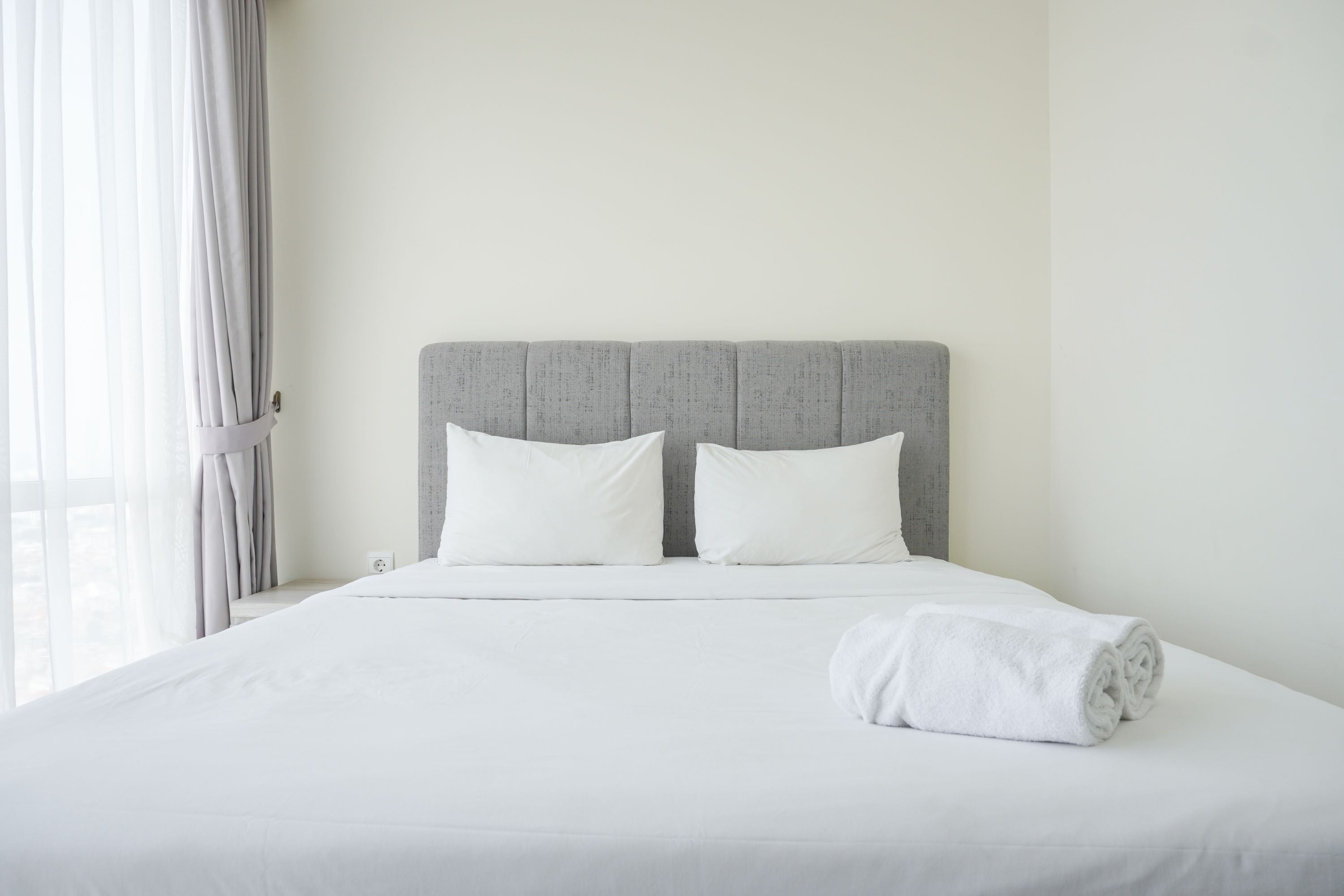 A neatly made bed with white linens and pillows, a grey upholstered headboard, and a white towel on the bed.