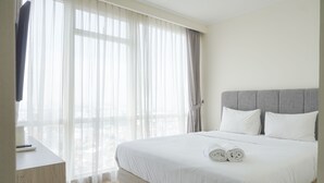 Ein ordentlich bezogenes Bett mit weißen Leinen und zusammengerollten Handtüchern, ein grauer möbelbezogener Kopfteil und leichte Vorhänge an einem Fenster.