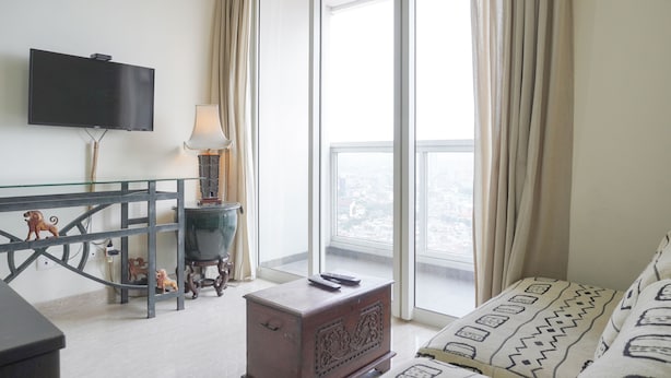 A hotel room with a flat-screen TV mounted on the wall, a glass coffee table, a wooden side table with a lamp, a small wooden chest, and a sofa with patterned cushions. - Nice And Elegant 2Br At 26Th Floor Menteng Park Apartment (Jakarta)