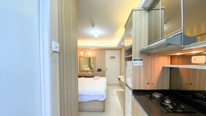 A compact hotel room with a kitchenette, a bed, a mirror, and a door leading to another room. - Cozy And Comfy Studio At Bassura City Apartment (Jakarta)