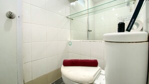 A bathroom with white tiles, a glass shower enclosure, a toilet with a red towel, and a glass shelf with toiletries. - Cozy And Comfy Studio At Bassura City Apartment (Jakarta)