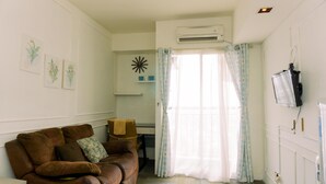 A living room with a brown sofa, a flat-screen TV mounted on the wall, and a window with light curtains.