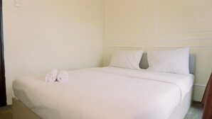 A neatly made bed with white linens and two rolled towels, a plain wall with a light switch, and a simple headboard.